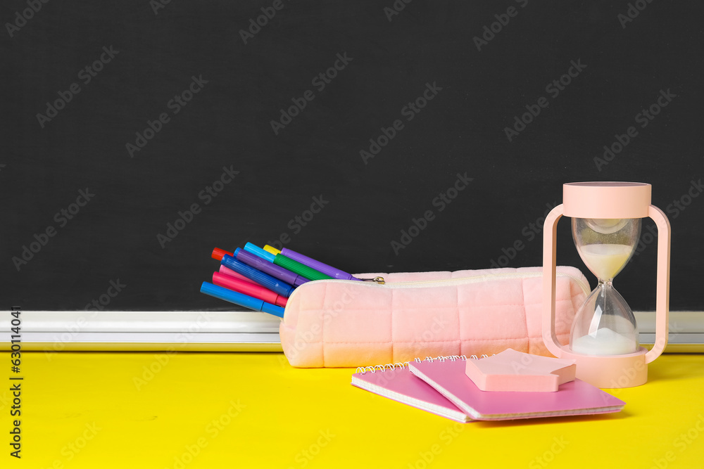 Pencil case with different school stationery and hourglass on yellow table near blackboard