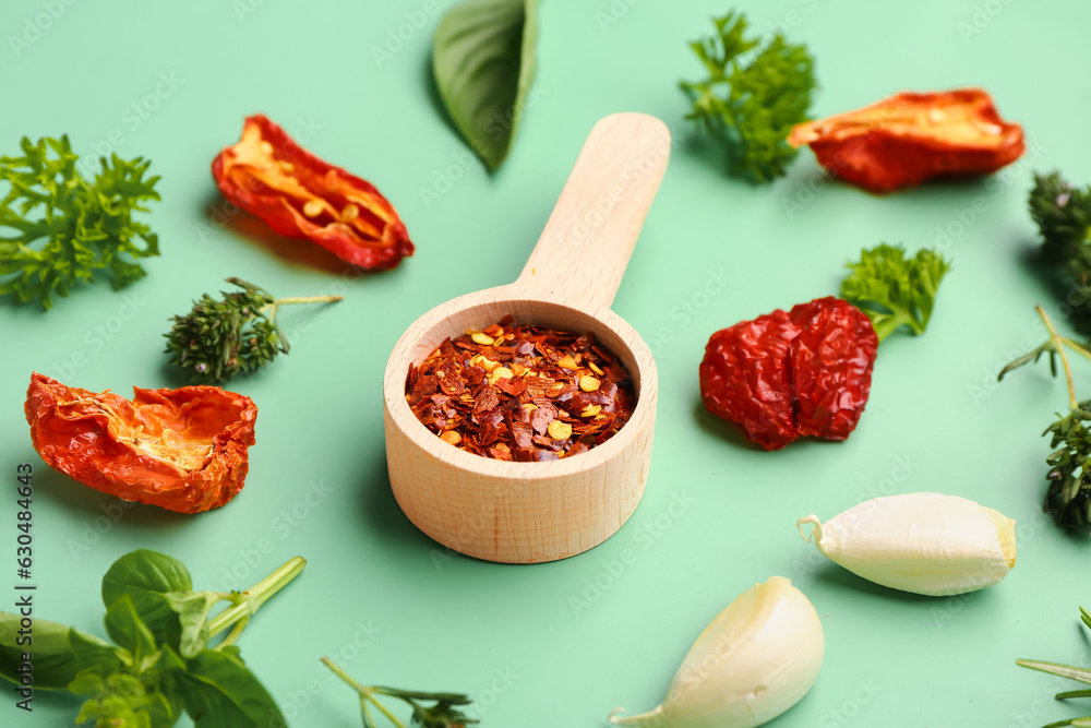 Wooden scoop with chili flakes, dried tomatoes and herbs on color background