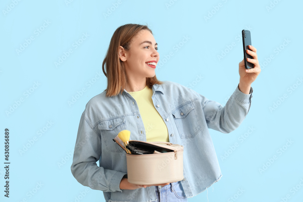 Young woman with cosmetic bag and mobile phone taking selfie on blue background