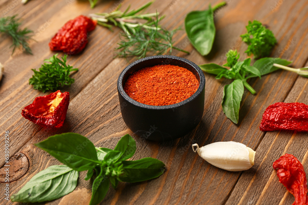 Bowl of paprika powder, dried tomatoes and herbs on wooden background