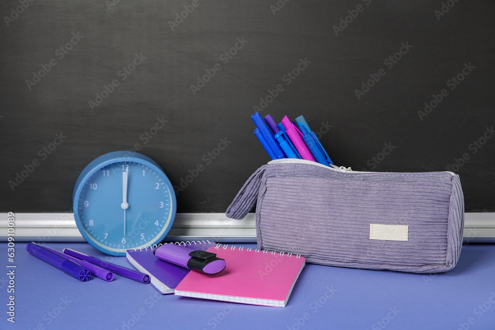 Pencil case with different school stationery and alarm clock on purple table near blackboard