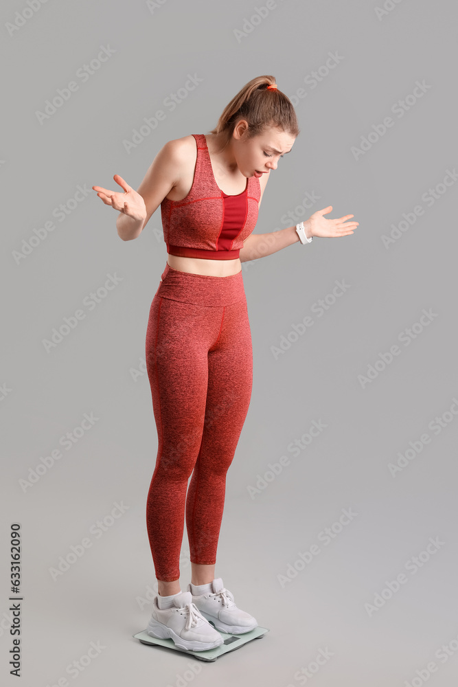 Confused young woman measuring her weight on scales against grey background