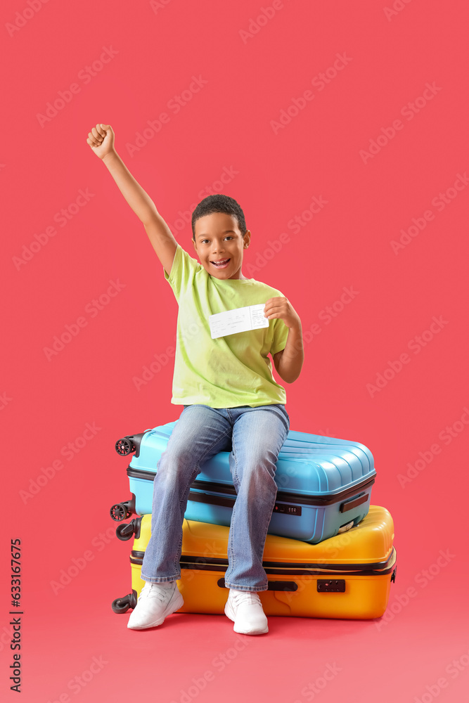 Happy African-American boy with ticket sitting on suitcases against red background