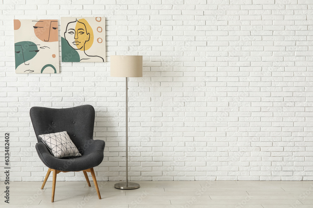 Grey armchair and lamp near white brick wall with different paintings