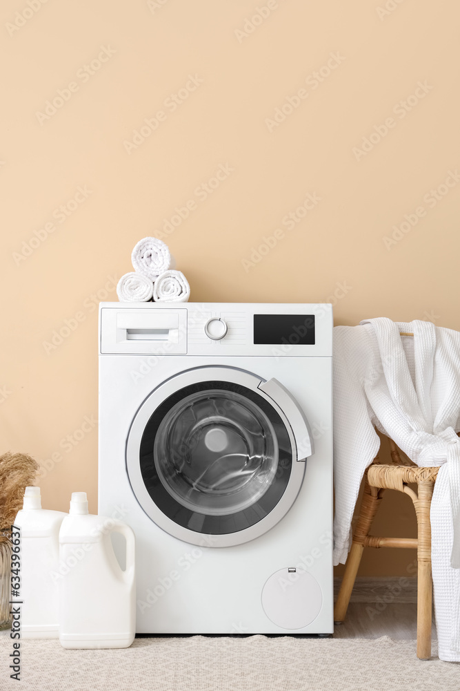 Washing machine, stool with bathrobe and bottles of detergent near beige wall