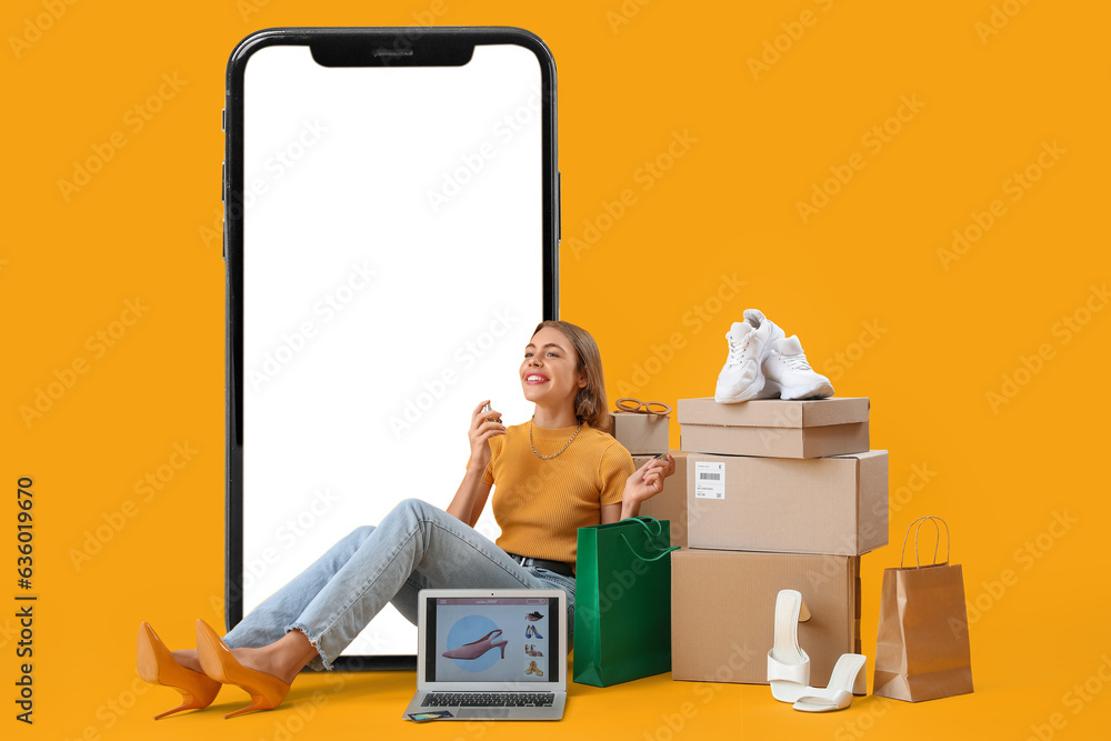 Stylish woman with new purchases and laptop sitting near big mobile phone on yellow background