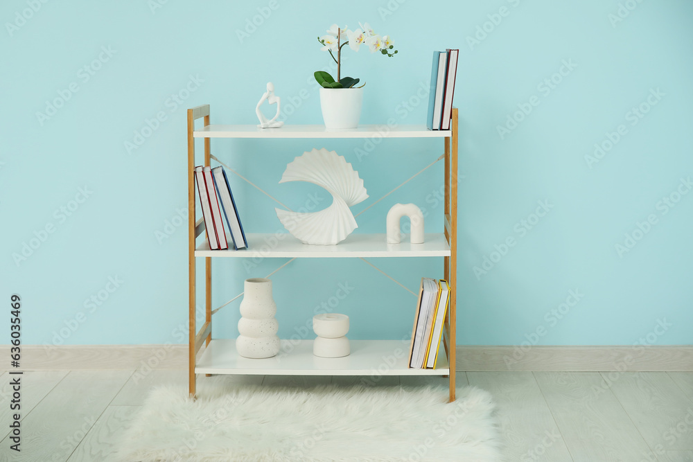 Shelving unit with books, orchid flower and decor near blue wall in room