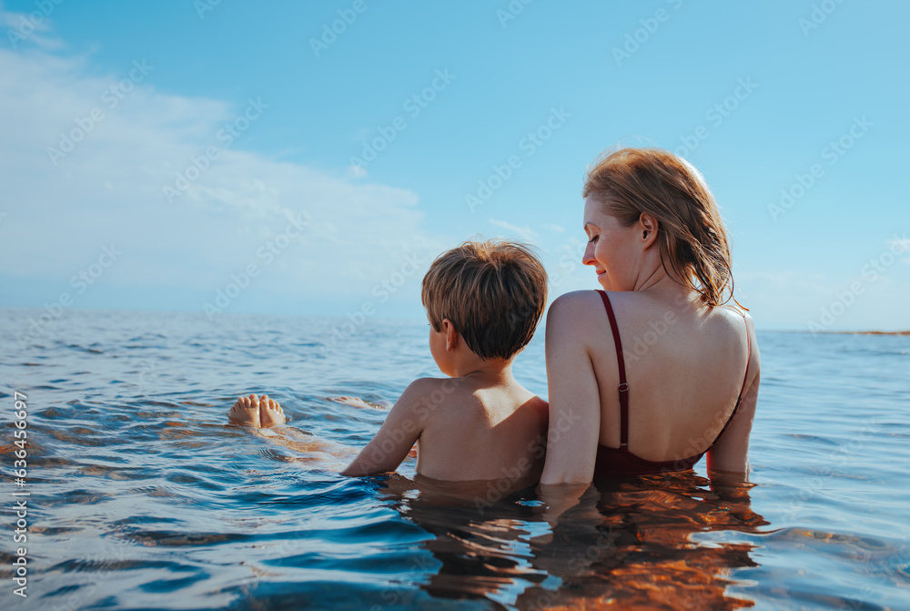 Mother and son sitting on seashore in water and looking away