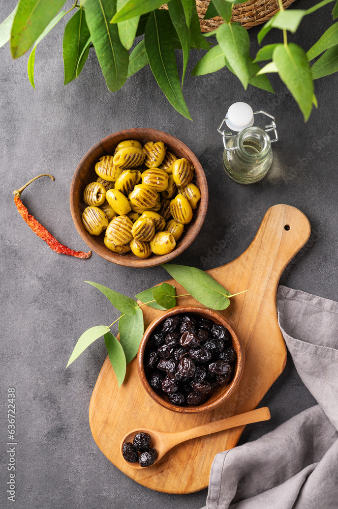 A set of green and black dried olives in bowls on a wooden cutting board on a dark background with o
