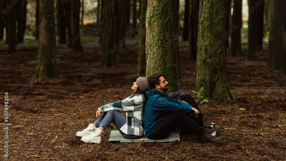 Tired cheerful young caucasian couple in jackets sits in forest on mat, enjoy travel at weekend, res