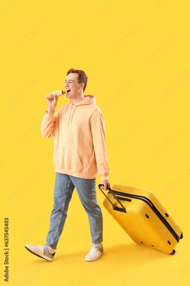 Going young man with tasty hot dog and suitcase on yellow background