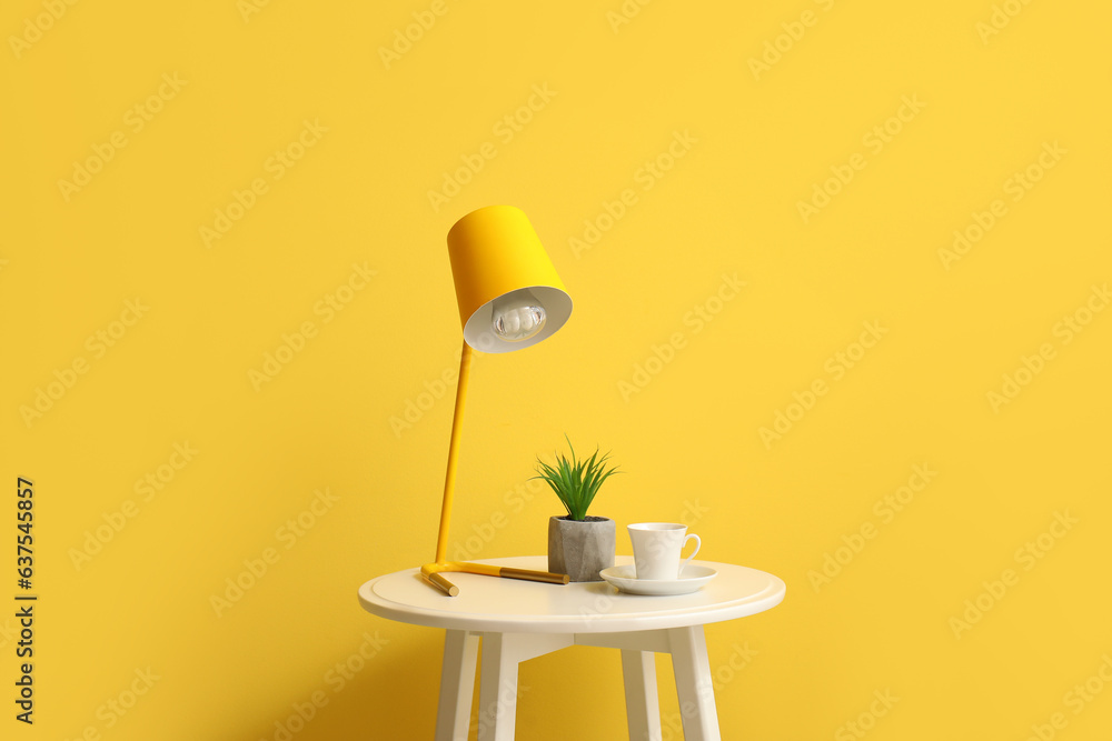 Small table with lamp, cup and houseplant near yellow wall