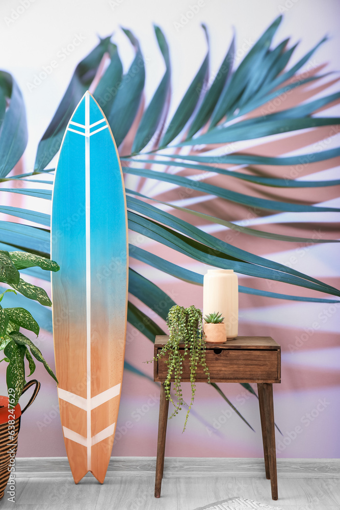 Wooden surfboard with table and houseplants near wall with printed tropical leaf