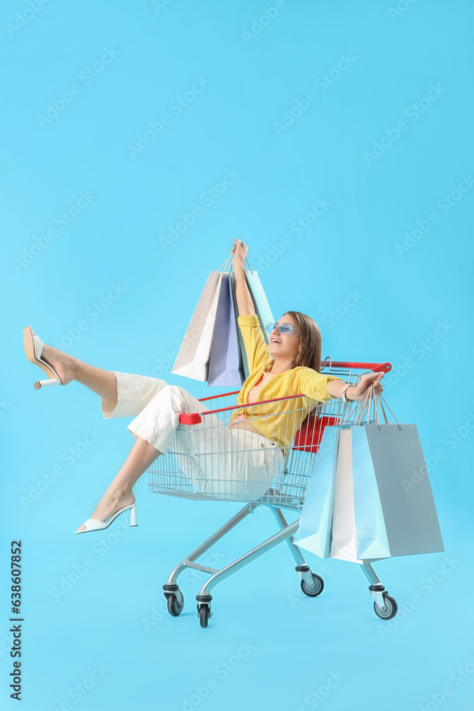 Stylish happy young woman with bags sitting in shopping cart on blue background