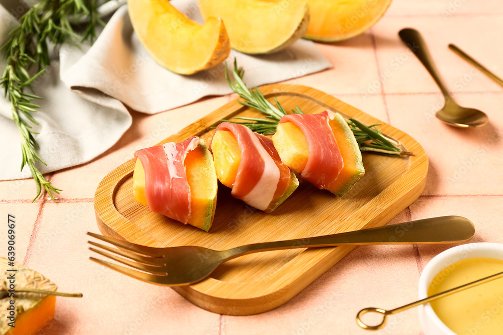 Wooden board of delicious melon with prosciutto and rosemary on pink tile background