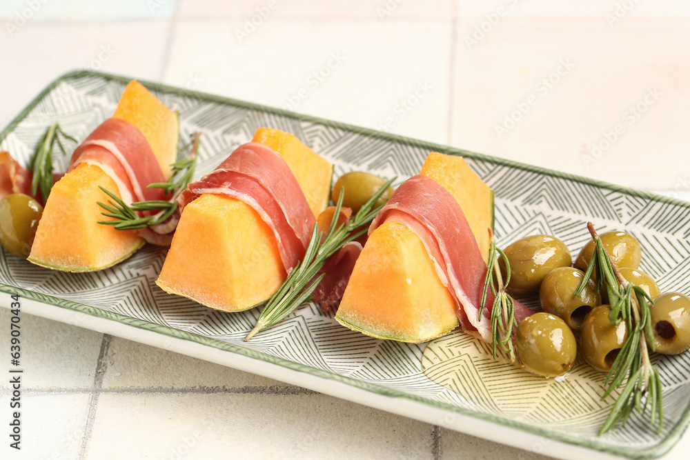 Plate with tasty melon, prosciutto, rosemary and green olives on light tile background, closeup