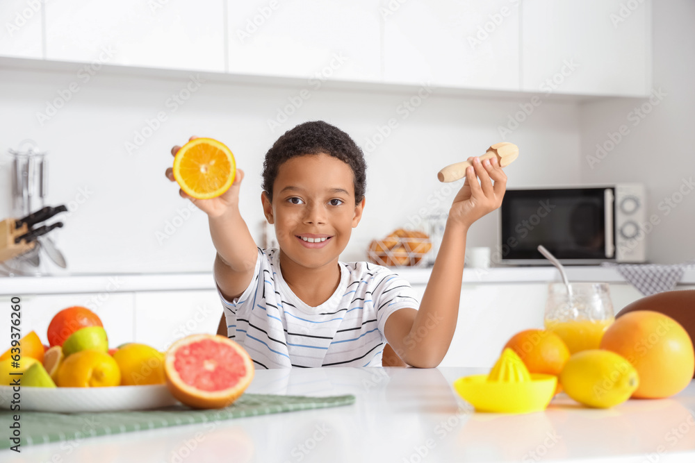 Little African-American boy with juicer and plate of different fresh citruses sitting at table in ki