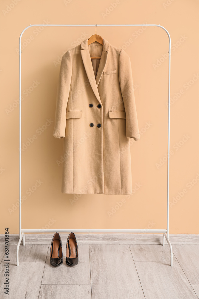 Stylish coat hanging on rack and high heeled shoes against color wall