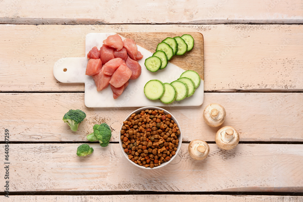 Composition with dry pet food, raw meat and natural products on light wooden background