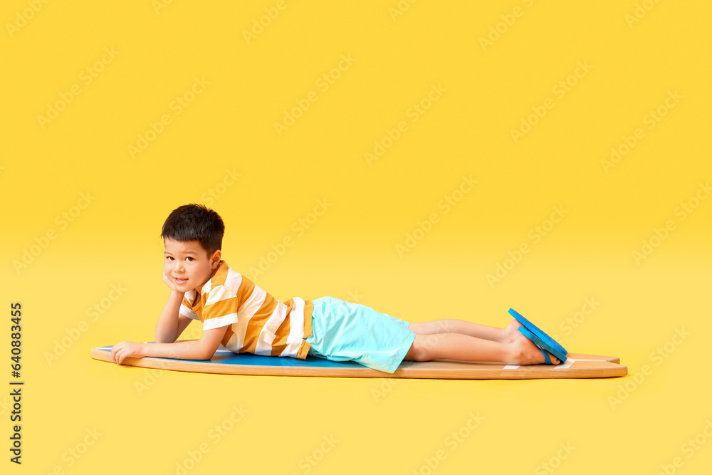 Cute little Asian boy lying on surfboard against yellow background