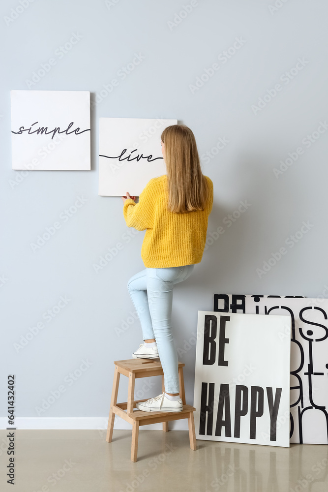 Young woman hanging poster on light wall at home