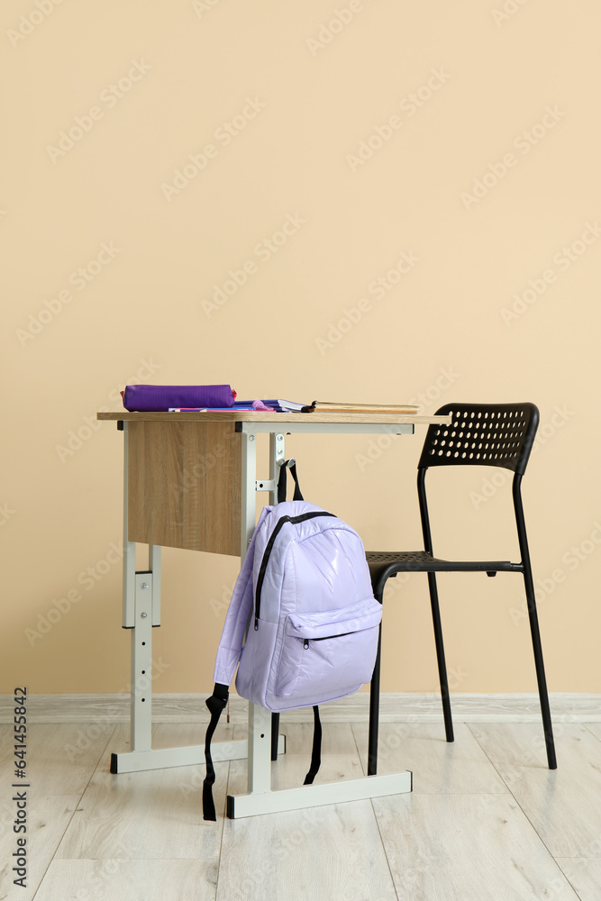 Modern school desk with backpack and stationery in room near beige wall