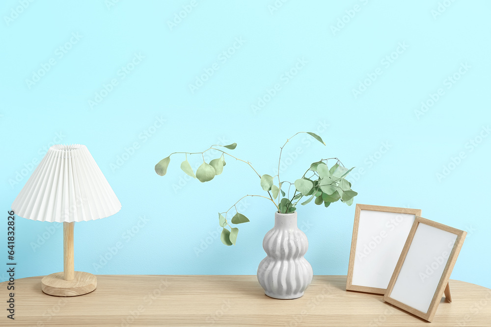 Vase of dried eucalyptus with lamp and photo frames on shelving unit near blue wall