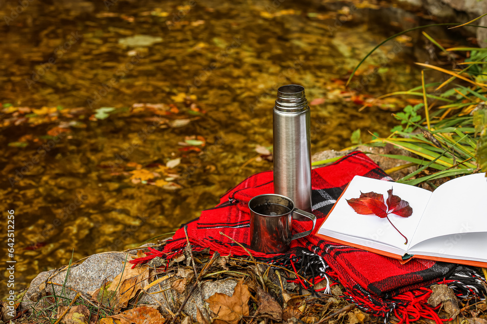 Book with autumn leaves, thermos, cup of tea and plaid near pond in park