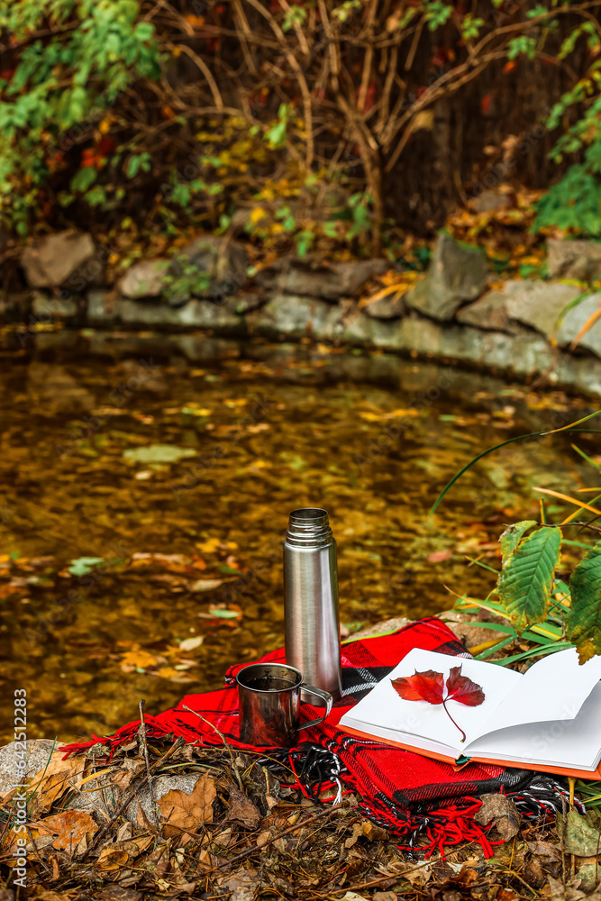 Book with autumn leaves, thermos, cup of tea and plaid near pond in park