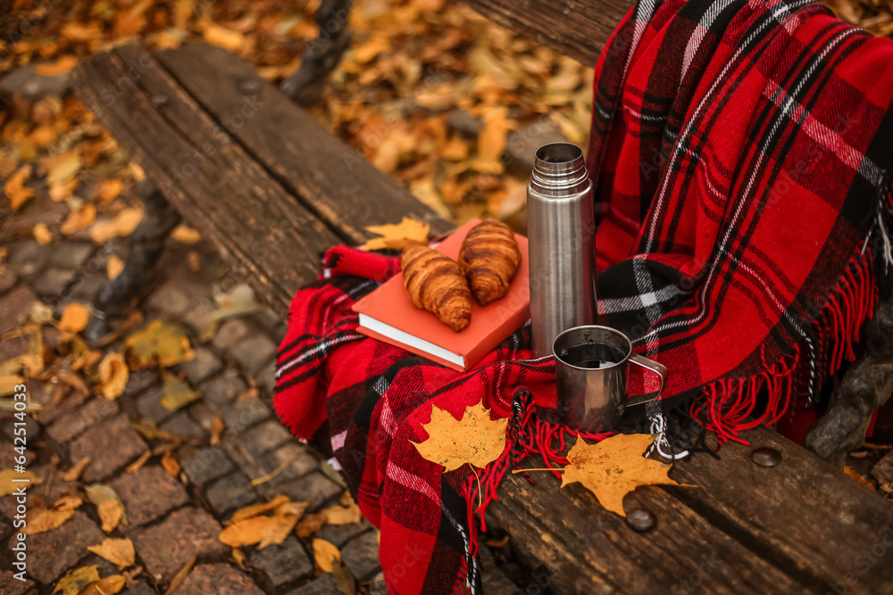 Book with croissants, thermos, cup of tea and plaid on bench in autumn park