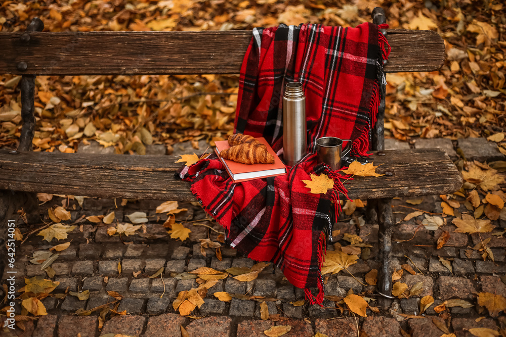 Book with croissants, thermos, cup of tea and plaid on bench in autumn park