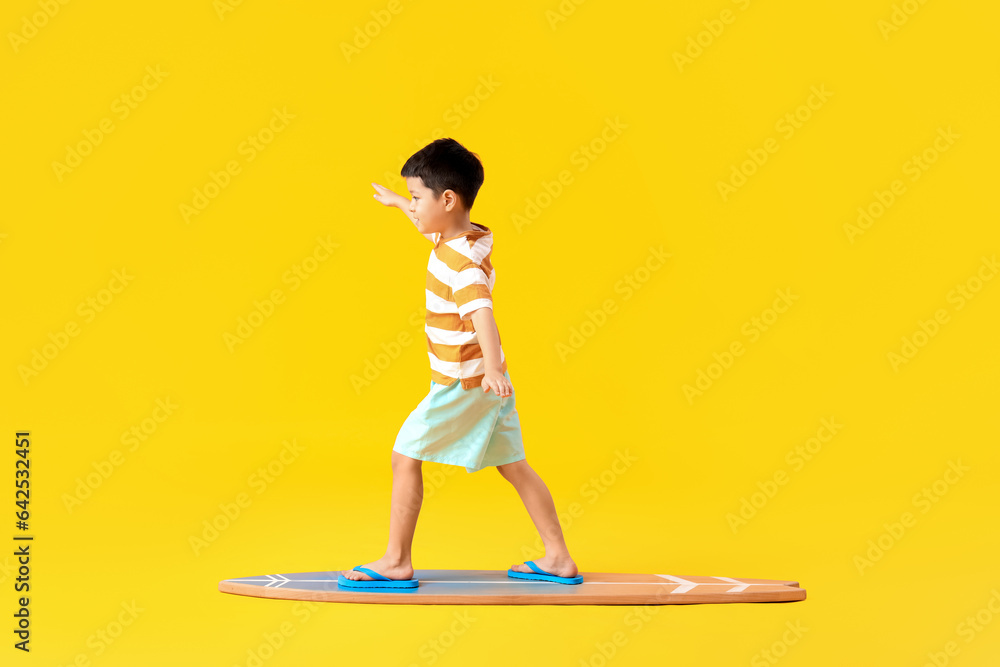 Cute little Asian boy standing on surfboard against yellow background