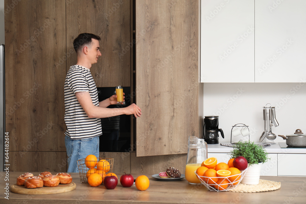 Young man with glass of orange juice opening cupboard in kitchen