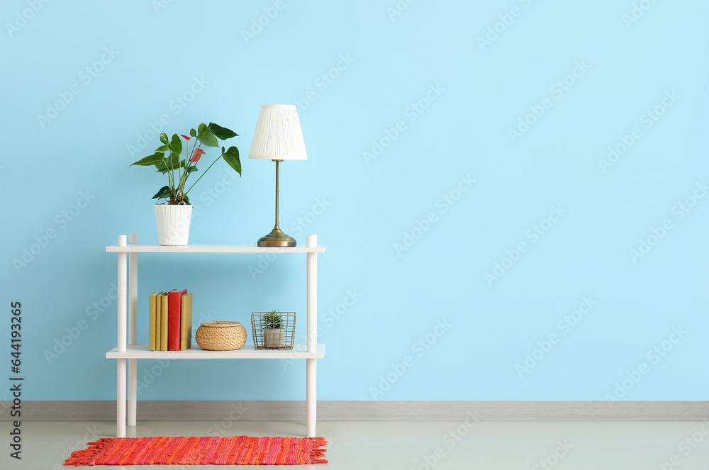 Shelving unit with houseplants, lamp and books near blue wall in room