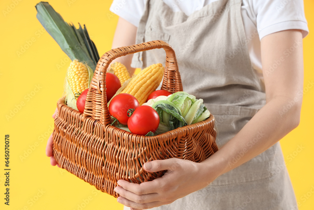 Young female farmer with wicker basket full of different ripe vegetables on yellow background, close