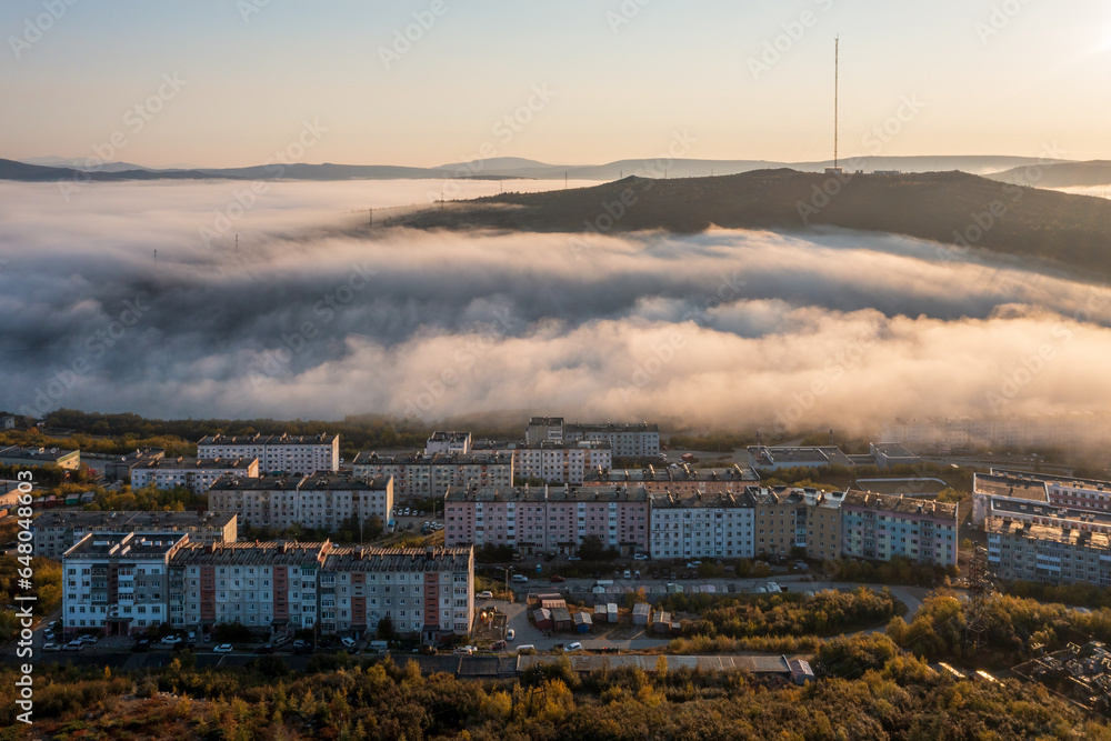 Amazing morning cityscape. Aerial view of residential buildings in the valley among the hills. Low clouds. TV tower on the top of the mountain. City of Magadan, Magadan region, Far East of Russia.