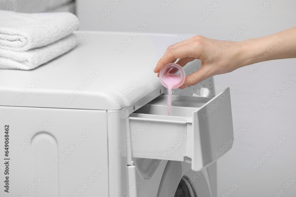Woman pouring fabric softener from cap into washing machine on light grey background, closeup