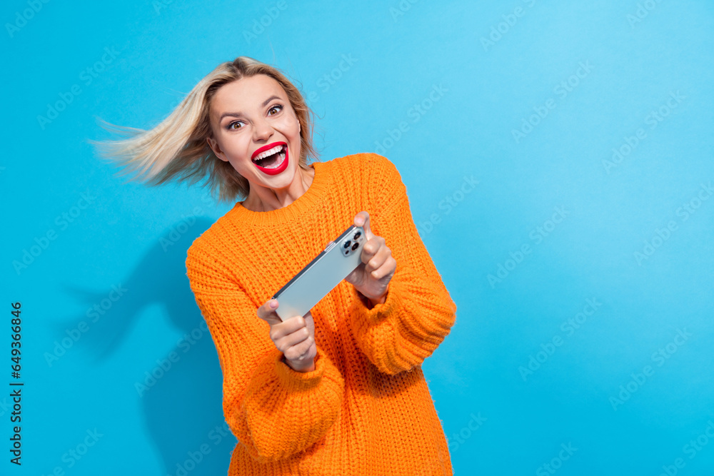Portrait of impressed woman fluttering hair dressed knitwear sweater hold smartphone play excited game isolated on blue color background