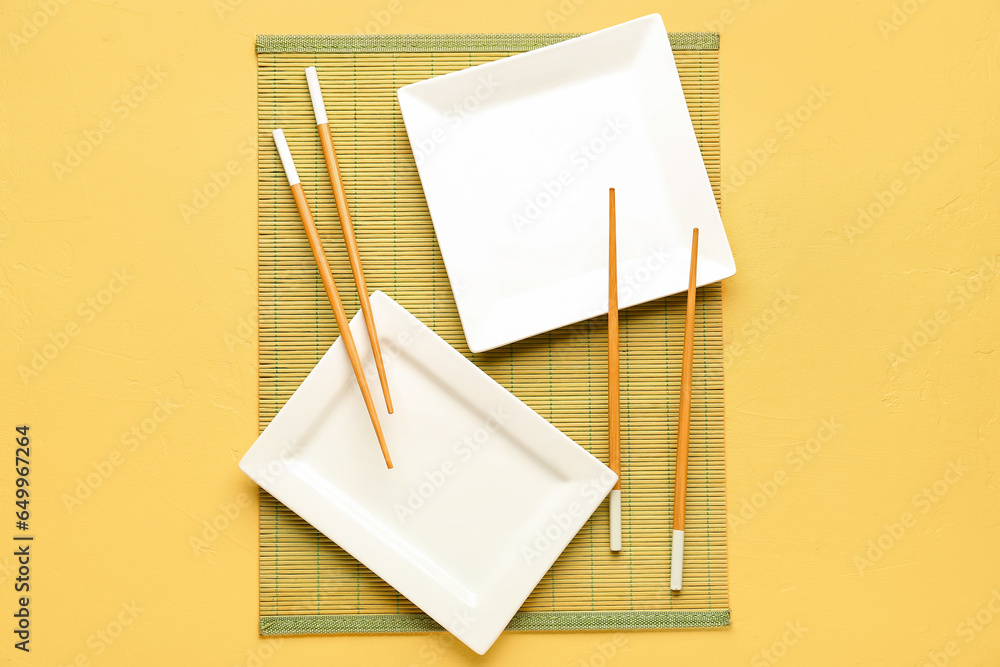 Chinese chopsticks and plates with bamboo mat on yellow background