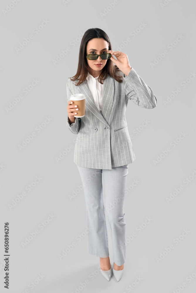 Young woman in stylish suit with cup of coffee on light background