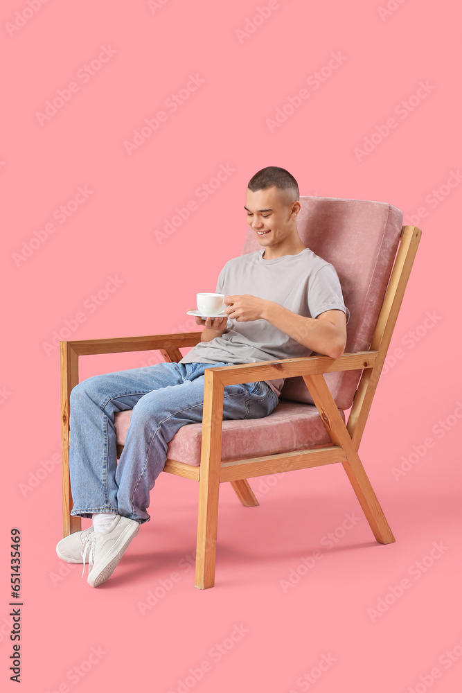Happy young man sitting on chair and drinking coffee against pink background