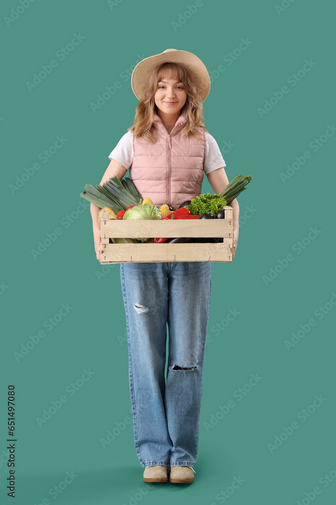 Happy young female farmer with wooden box full of different ripe vegetables on green background