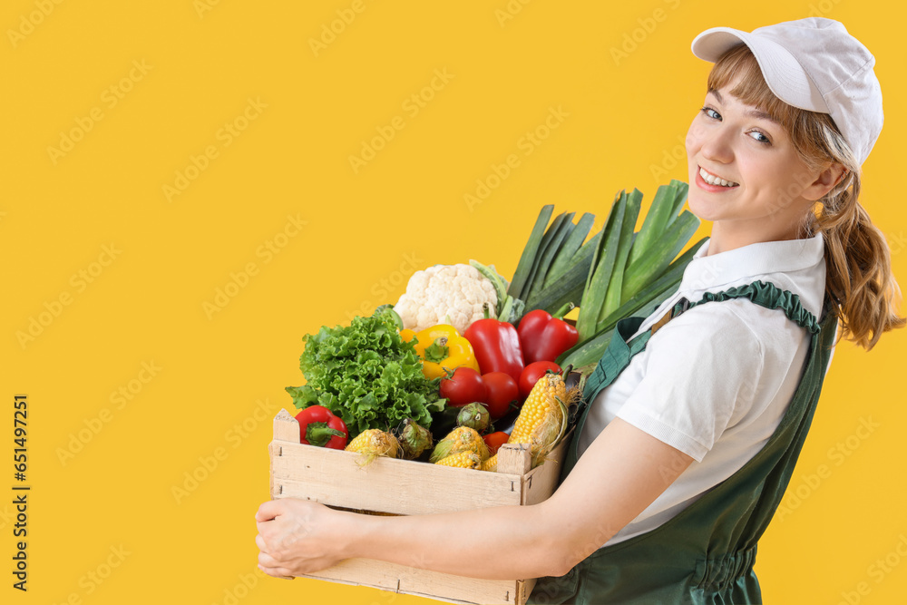 Happy young female farmer with wooden box full of different ripe vegetables on yellow background