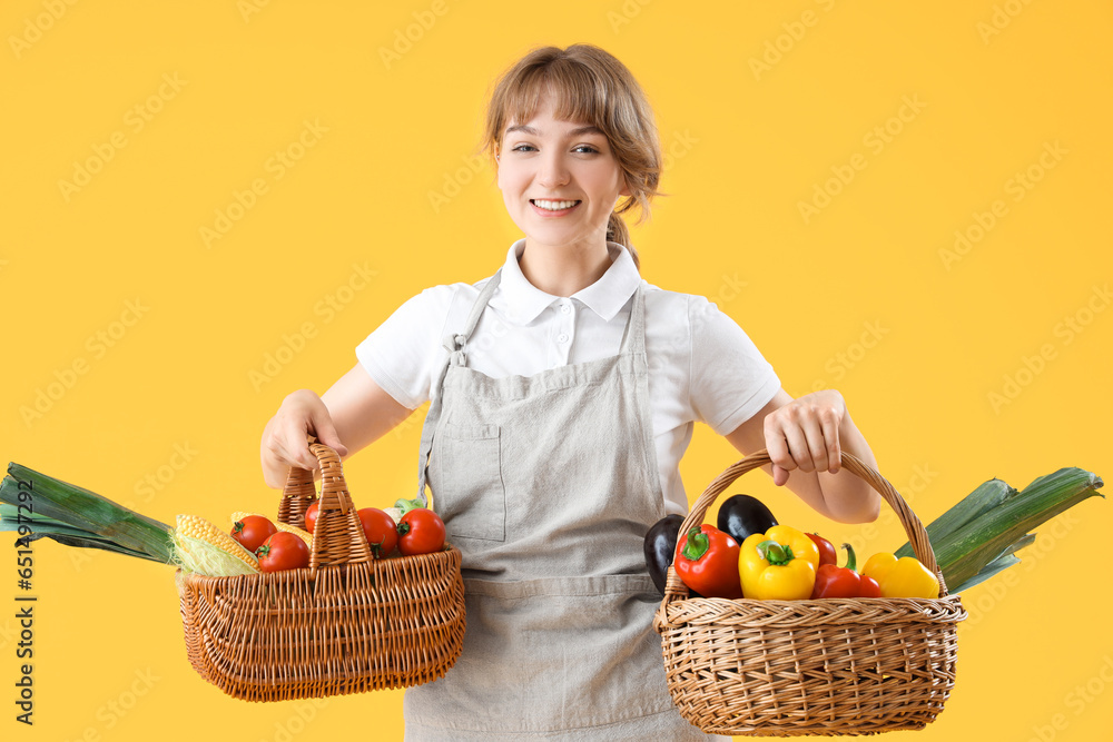 Happy young female farmer with wicker baskets full of different ripe vegetables on yellow background