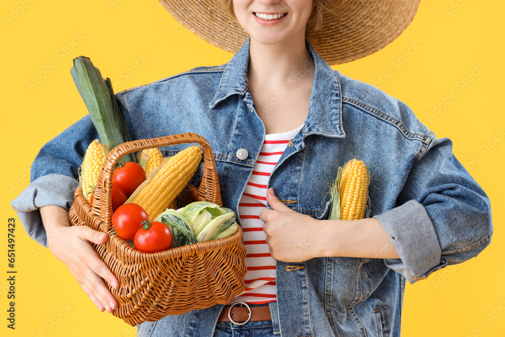 Young female farmer with corn and wicker basket full of different ripe vegetables on yellow background, closeup