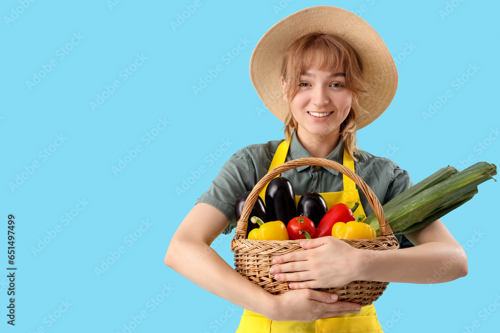 Happy young female farmer with wicker basket full of different ripe vegetables on blue background