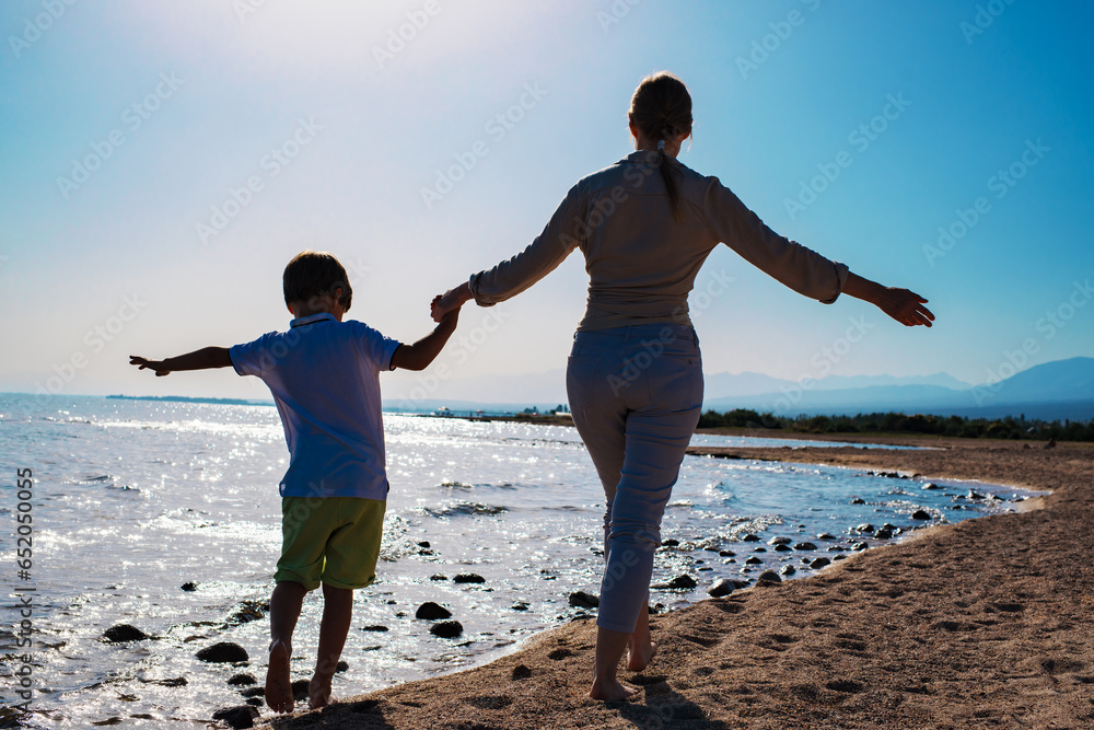 Young woman and her son walking barefoot on sandy beach holding hands