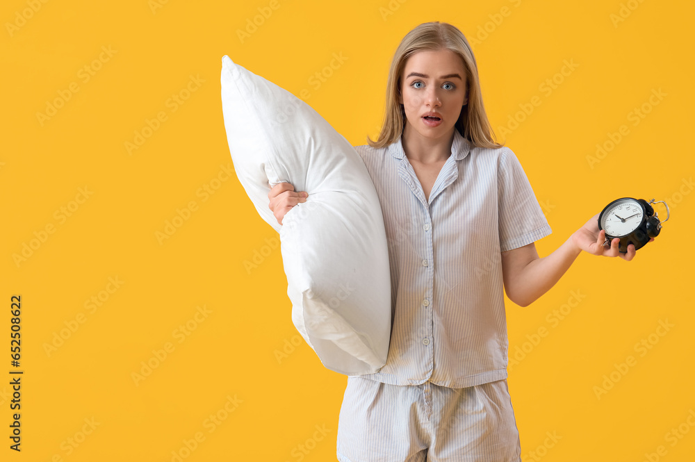 Confused young woman with pillow and alarm clock on yellow background