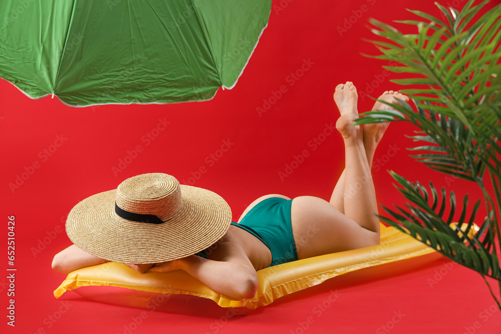 Young beautiful woman in wicker hat with umbrella lying on inflatable mattress against red background. Summer vacation concept