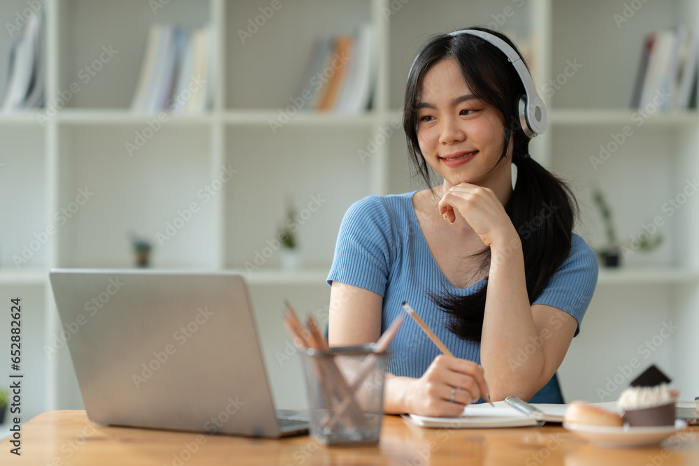 Asian woman wearing headphones sitting and working Listen to relaxing music and chat with friends on social media on laptop while sitting on weekends home office desk.